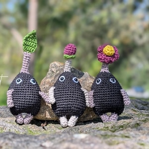 May include: Three handmade crochet creatures with black bodies, white eyes, and small arms and feet. Each has a different flower or leaf on top, including a green leaf, a purple bud, and a pink flower with a yellow center. The creatures are displayed on a rock.