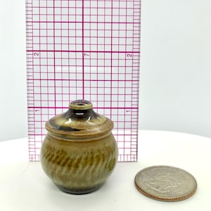 May include: A small, handmade ceramic jar with a lid. The jar is approximately 2 inches tall and has a brown and green glaze with a textured pattern. A quarter is next to the jar for size comparison.