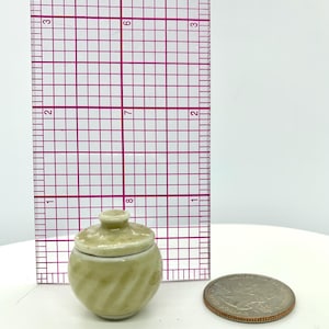 May include: A small, light green ceramic jar with a lid, featuring a spiral design. The jar is next to a ruler and a quarter for size comparison. The jar is approximately 1 inch tall.