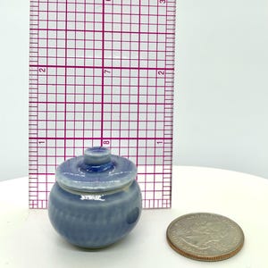 Może przedstawiać: Mały, jasnoniebieski ceramiczny słoik z pokrywką, obok linijki i monety. Słoik ma okrągły korpus z pionowymi liniami i małym guzikiem na pokrywce. Słoik ma około 2,5 cm wysokości.