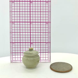 Puede incluir: Un pequeño tarro de cerámica verde claro con tapa, junto a una regla y una moneda. El tarro mide aproximadamente 2,5 cm de alto. El tarro tiene un cuerpo redondeado y una tapa pequeña y redondeada. La moneda es plateada y tiene un borde marrón.