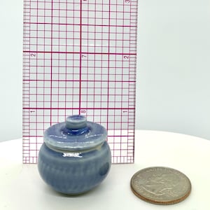 May include: A small, light blue ceramic jar with a lid, next to a ruler and a quarter. The jar has a round body with vertical lines and a small knob on the lid. The jar is approximately 1 inch tall.