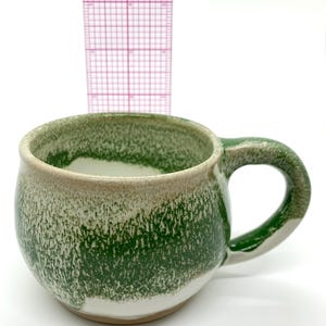 Può includere: Una tazza in ceramica fatta a mano con forma arrotondata e un grande manico. La tazza presenta una glassa verde macchiettata su una base bianca, con un bordo beige. La tazza è alta circa 9 cm.