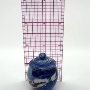 Puede incluir: Un pequeño tarro de cerámica azul con tapa. El tarro mide aproximadamente 5 cm de alto y presenta un diseño pintado a mano con detalles blancos y rojos. El tarro tiene forma redonda y un pequeño pomo en la tapa.