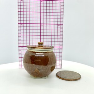 May include: A small, brown ceramic jar with a lid. The jar has a rounded shape and a textured surface. The lid is a lighter brown color with a small knob on top. A ruler is in the background. A quarter is next to the jar.