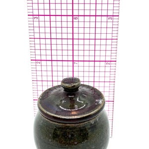 Op de afbeelding: Een kleine, handgemaakte keramische pot met een deksel. De pot is donkergroen met bruine en paarse accenten. De pot is ongeveer 7.6 cm hoog.