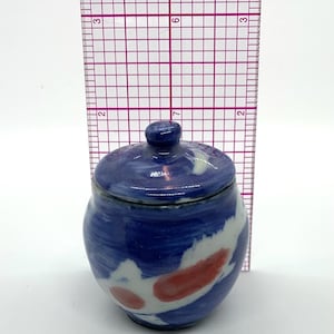 Puede incluir: Un pequeño tarro de cerámica azul con tapa. El tarro mide aproximadamente 7.6 cm de alto y presenta diseños abstractos blancos y rojos. Tiene forma redondeada y un pequeño pomo en la tapa. Probablemente hecho a mano, es adecuado para guardar objetos pequeños.
