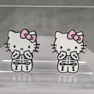 May include: Two white Hello Kitty-shaped earrings with pink bows. Each earring features a black skeleton design on the lower half. The earrings are displayed on a clear, reflective surface.