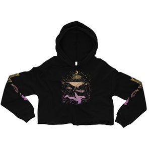 May include: Black cropped hoodie featuring a graphic of a whale, orcas, and fish within a geometric shape adorned with stars and a crescent moon. The graphic is rendered in shades of pink and gold.
