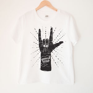 May include: White t-shirt with a black and white illustration of a hand making the 'I love you' sign. The hand has a tattoo that says 'Mother Earth' with a heart.