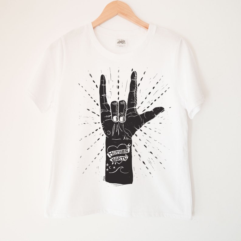 May include: White t-shirt with a black and white illustration of a hand making the 'I love you' sign. The hand has a tattoo that says 'Mother Earth' with a heart.
