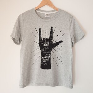 May include: A grey t-shirt with a black graphic of a hand making the 'I love you' sign. The hand has a tattoo that says 'Mother Earth' with a heart.