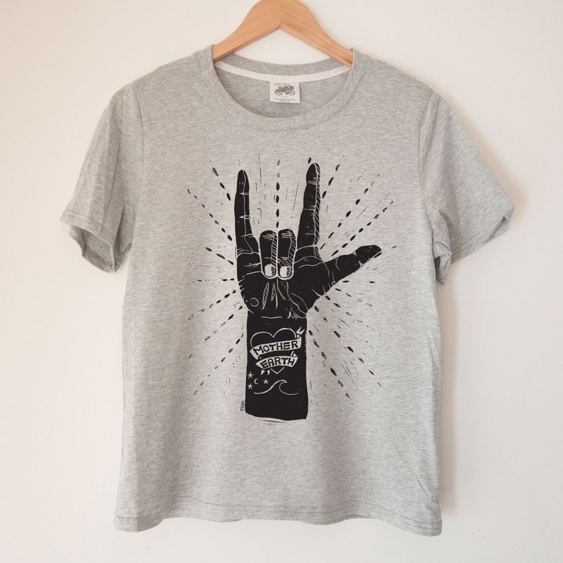 May include: A grey t-shirt with a black graphic of a hand making the 'I love you' sign. The hand has a tattoo that says 'Mother Earth' with a heart.