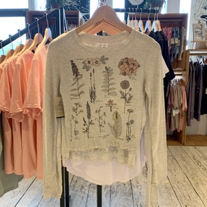 May include: A light grey long-sleeved shirt with a floral print featuring various types of flowers and plants. The shirt has a white underlayer and a ruffled hem.