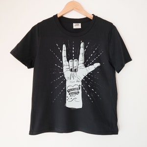 May include: Black t-shirt with a white graphic of a hand making the 'I love you' sign with the words 'Mother Earth' on the wrist.