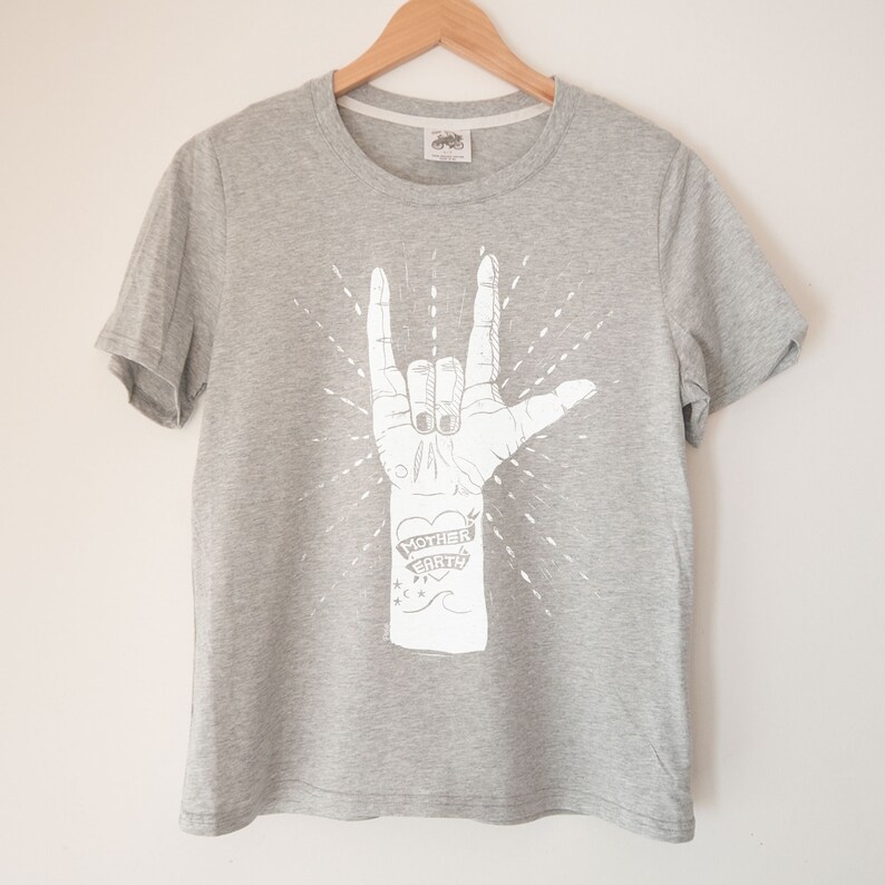 May include: Gray t-shirt with a white graphic of a hand making the 'I love you' sign with the words 'Mother Earth' on the wrist.