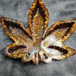 May include: A resin ashtray shaped like a cannabis leaf, with a brown and gold glitter finish. The leaf design includes detailed veins and a small, raised rest area. The ashtray is designed for use.