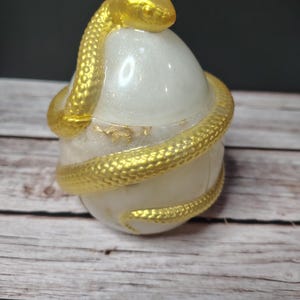 Può includere: Un contenitore bianco perlato a forma di uovo con coperchio rimovibile, caratterizzato da un serpente dorato metallizzato e testurizzato avvolto attorno all'esterno. La base include scaglie di foglia d'oro e la testa del serpente poggia in cima.