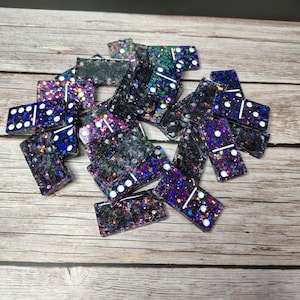 May include: A set of rectangular dominoes made from dark resin with embedded multi-coloured glitter and holographic flecks. Each piece features white painted pips and lines indicating numerical values, scattered in a pile on a wooden surface.
