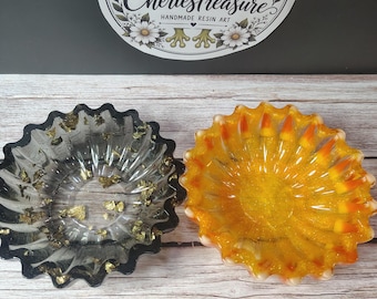Puede incluir: Dos cuencos de resina festoneados. El cuenco izquierdo es negro translúcido con copos de oro incrustados. El cuenco derecho es naranja y amarillo translúcido con trozos de maíz dulce incrustados. Arriba hay un logotipo con una rana y el texto CheriesTreasure.
