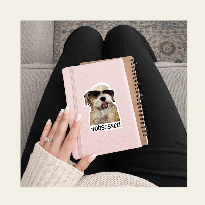 A person sits on a couch with a pink notebook on their lap, decorated with a custom dog sticker featuring a pup in sunglasses and the text “#obsessed.” The person wears black pants and a cream sweater, with manicured nails and a ring visible. A patterned rug and couch upholstery complete the cozy scene, showcasing how custom pet stickers personalize journals and everyday items.