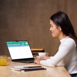 May include: A woman is seated at a wooden table, using a laptop. The laptop screen displays a spreadsheet labeled "Stash Inventory Kit." A glass of orange juice and a cup of coffee are on the table. Notebooks are stacked in front of the laptop.