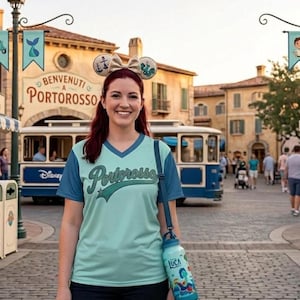 May include: A woman wearing a light blue and teal V-neck shirt with "Portorosso" text, a matching water bottle, and mouse ears. The water bottle features a cartoon design and the name "Luca". The background shows a street scene with buildings and a trolley.