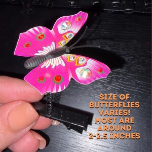 Op de afbeelding: Een roze vlinderhaarclip met zwarte voelsprieten en een zwarte veerbasis. De vleugels hebben rode en iriserende cirkelvormige accenten. De tekst luidt: SIZE OF BUTTERFLIES VARIES! MOST ARE AROUND 2-2.5 INCHES.