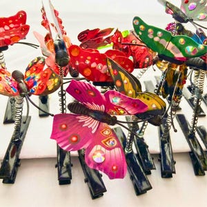 Op de afbeelding: Een verzameling decoratieve vlinderclips met levendig gekleurde vleugels in rood, roze, groen en geel. Elke vlinder is versierd met iriserende strass-steentjes en gemonteerd op een flexibele metalen veer die aan een zwarte krokodillenklem is bevestigd.