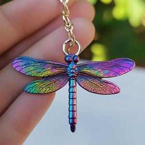 Peut inclure: Un pendentif libellule coloré avec des ailes irisées dans les tons de violet, bleu et vert. La libellule est suspendue à une chaîne dorée. Les ailes présentent des motifs de veines détaillés.