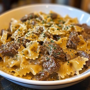 May include: A close-up of a white bowl filled with pasta, beef, and a creamy sauce. The pasta is bowtie shaped and the beef is in small chunks. The sauce is a light brown color and there are small green herbs sprinkled on top.