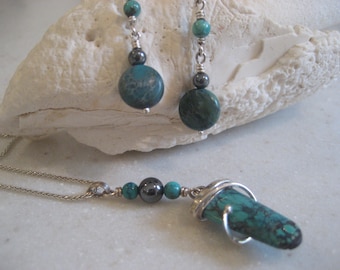 May include: A turquoise and silver jewelry set featuring a pendant necklace and matching earrings. The necklace has a silver chain with turquoise and dark gray beads. The earrings have turquoise and dark gray beads suspended from silver wires.