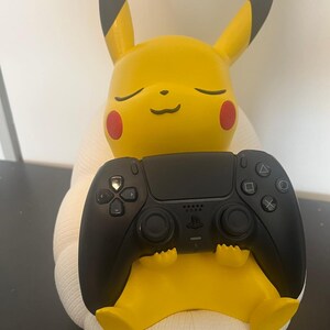 May include: A yellow Pikachu figure with pointed ears, red cheeks, and closed eyes, sitting on a white textured base. The figure holds a black wireless video game controller with a central touchpad, directional buttons, and action symbols.