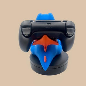 Puede incluir: Un mando de juego Sony negro descansa sobre un soporte azul y naranja con forma de personaje. El mando cuenta con botones superiores etiquetados como R2 y L2, con la marca Sony impresa en el borde superior, sobre una base negra circular.