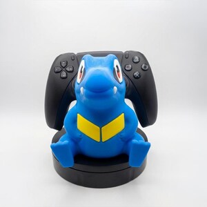 May include: A blue and yellow cartoon character controller holder with a black controller behind it. The holder sits on a black base. The controller has buttons and a directional pad.