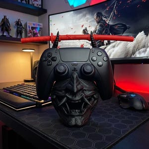 May include: A black PlayStation 5 controller rests on a black Oni mask-shaped stand, with a red katana-style controller holder. The stand is on a black hexagonal patterned mousepad. A computer monitor and keyboard are in the background.
