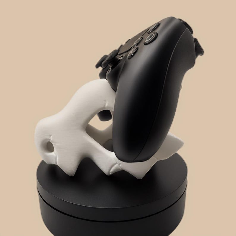 Puede incluir: Un mando de videojuegos negro apoyado sobre un soporte blanco de formas org&aacute;nicas y abstractas. El soporte presenta varios huecos y se asienta sobre una base circular negra s&oacute;lida, proporcionando un montaje estable para el perif&eacute;rico.
