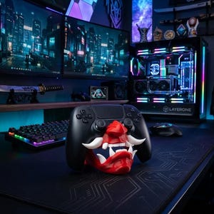 May include: A black PlayStation 5 controller rests on a red and white Oni mask stand. The stand is on a black mousepad with a circuit board design. A gaming PC with LED lights and multiple monitors displaying a cityscape are in the background.
