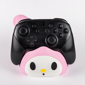 May include: A black Nintendo Switch Pro controller resting in a pink and white character-themed stand. The stand features a rounded face with two black oval eyes, a small yellow nose, and a tiny black mouth, resembling a cartoon character.