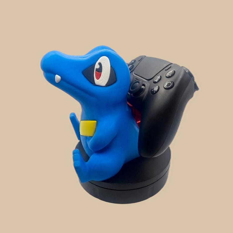 Puede incluir: Una figura de personaje azul de tipo reptil con un parche amarillo en el pecho, ojos grandes blancos y rojos y un solo diente blanco, montada sobre una base circular negra y sosteniendo un mando de videojuego negro en su espalda.
