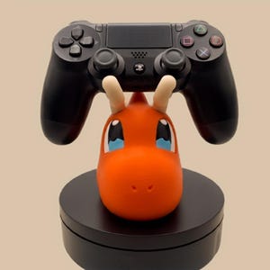 Puede incluir: Un mando de videojuego negro descansa sobre un soporte con forma de cabeza de drag&oacute;n naranja con cuernos color crema y grandes ojos azules y blancos. La cabeza del personaje est&aacute; montada sobre una base circular negra s&oacute;lida que sostiene el mando.