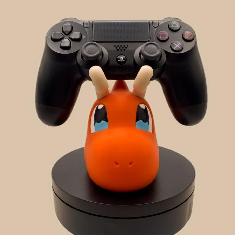 Puede incluir: Un mando de videojuego negro descansa sobre un soporte con forma de cabeza de drag&oacute;n naranja con cuernos color crema y grandes ojos azules y blancos. La cabeza del personaje est&aacute; montada sobre una base circular negra s&oacute;lida que sostiene el mando.