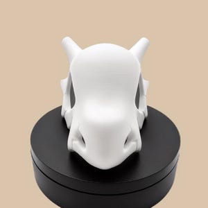 Puede incluir: Una figura de color blanco mate impresa en 3D con forma de cr&aacute;neo, que presenta dos protuberancias en forma de cuerno en los lados superiores, un cr&aacute;neo redondeado y liso, y una cavidad nasal definida, sobre una base circular negra.