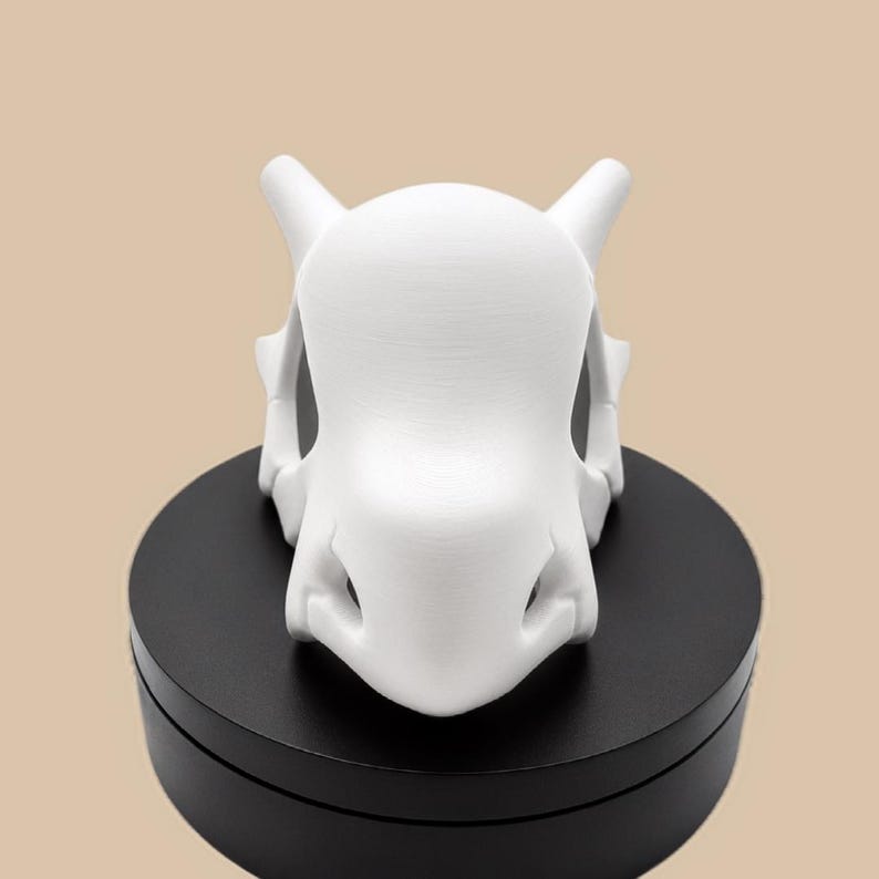 Puede incluir: Una figura de color blanco mate impresa en 3D con forma de cr&aacute;neo, que presenta dos protuberancias en forma de cuerno en los lados superiores, un cr&aacute;neo redondeado y liso, y una cavidad nasal definida, sobre una base circular negra.