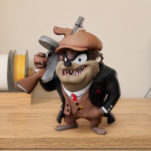 May include: A stylized figurine of the Tasmanian Devil dressed as a 1920s gangster, wearing a brown flat cap, a black overcoat, and a tan vest with a red tie. The character holds a grey and brown toy submachine gun over its right shoulder.
