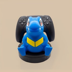 Puede incluir: Un soporte para mando en forma de criatura azul y amarilla que sostiene un mando de juego negro. El soporte presenta una figura azul de estilo dibujo animado con ojos grandes, colmillos blancos y un patr&oacute;n de cheur&oacute;n amarillo sobre una base negra.