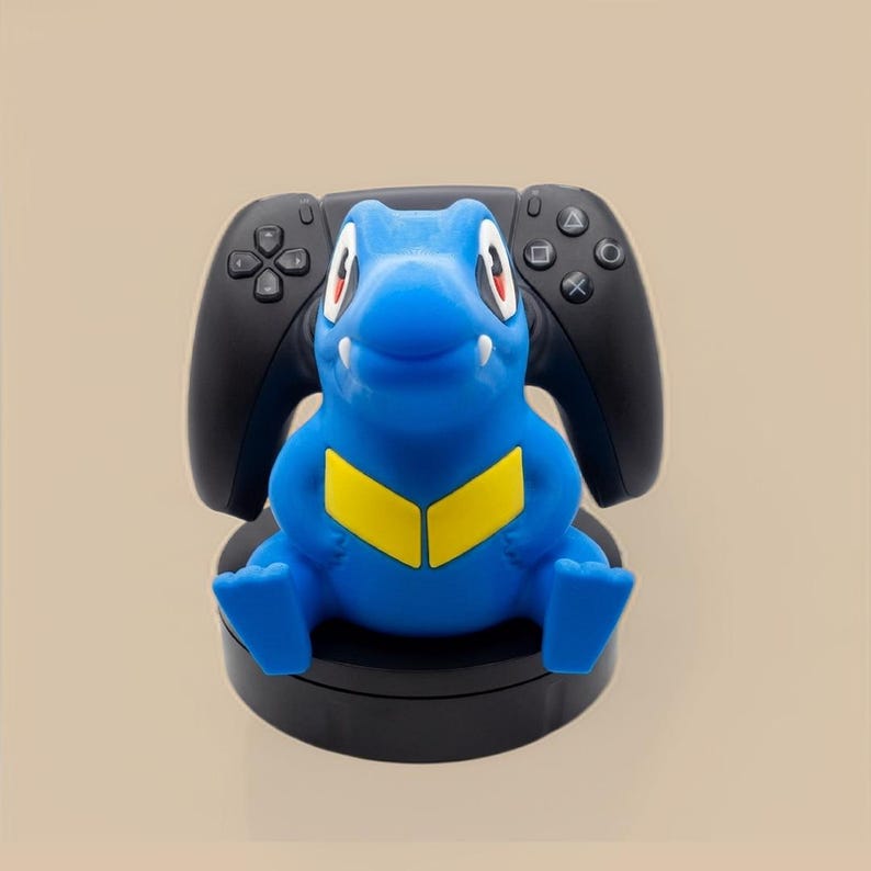 Puede incluir: Un soporte para mando en forma de criatura azul y amarilla que sostiene un mando de juego negro. El soporte presenta una figura azul de estilo dibujo animado con ojos grandes, colmillos blancos y un patr&oacute;n de cheur&oacute;n amarillo sobre una base negra.