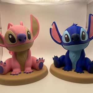 May include: Two character figurines sitting on tan oval bases. The left figure is light pink with tan accents and large ears. The right figure is dark blue with light blue accents and large ears. Both figures have large black eyes and purple noses.