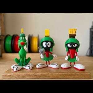 Puede incluir: Tres figuras de personajes de dibujos animados sobre una superficie de madera. Las figuras incluyen un personaje verde con forma de perro y dos versiones de un personaje con casco verde, torso rojo y guantes y zapatos blancos. Uno sostiene una pistola de rayos.
