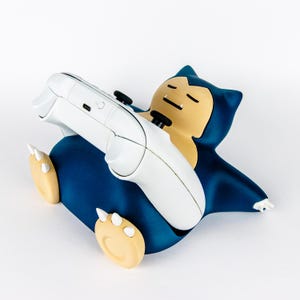 Peut inclure: Un support de manette en forme de Ronflex bleu foncé et beige tenant une manette de jeu vidéo sans fil blanche. Le personnage a une expression endormie avec les yeux fermés et de petites griffes blanches sur les pieds et les mains.